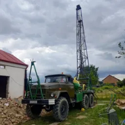 Wiertnica na podwoziu ciężarówki ZIL z rozłożonym masztem wiertniczym na tle zachmurzonego nieba i zabudowań gospodarczych w trakcie wiercenia studni.