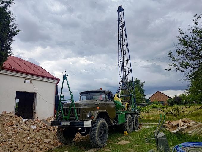 Wiertnica na podwoziu ciężarówki ZIL z rozłożonym masztem wiertniczym na tle zachmurzonego nieba i zabudowań gospodarczych w trakcie wiercenia studni.