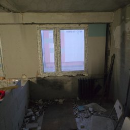 Adrian panfil - Wnętrze w trakcie prac przygotowawczych, widoczne okno z napisem 'ROMANTYCZNA', ściany z fragmentami płyt kartonowo-gipsowych, surowy strop i gruz na podłodze, krzesło z tworzywa sztucznego w rogu.