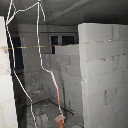 Adrian panfil - Wnętrze w trakcie budowy ścian działowych z białych bloczków, widoczne przewody elektryczne zwisające z sufitu i okno w tle.