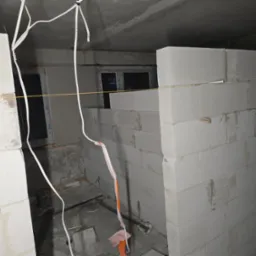 Wnętrze w trakcie budowy ścian działowych z białych bloczków, widoczne przewody elektryczne zwisające z sufitu i okno w tle.