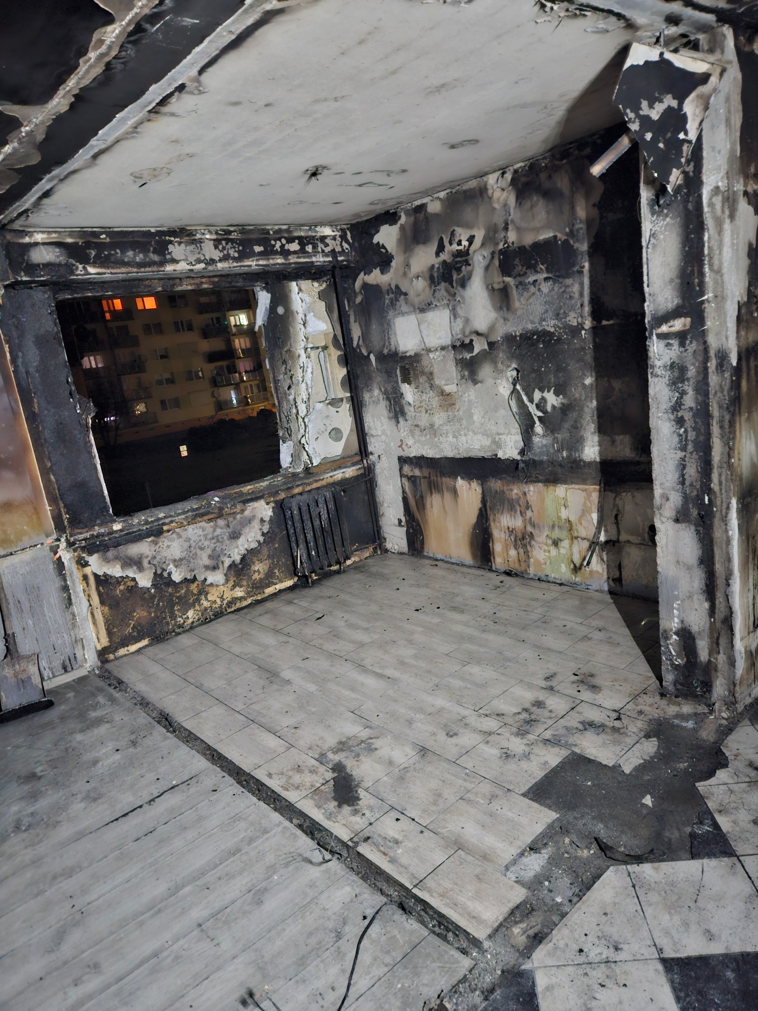 Wnętrze pokoju po pożarze: osmalone ściany, okopcona instalacja grzewcza, zniszczona podłoga z płytek, widok na oświetlony blok mieszkalny za oknem.