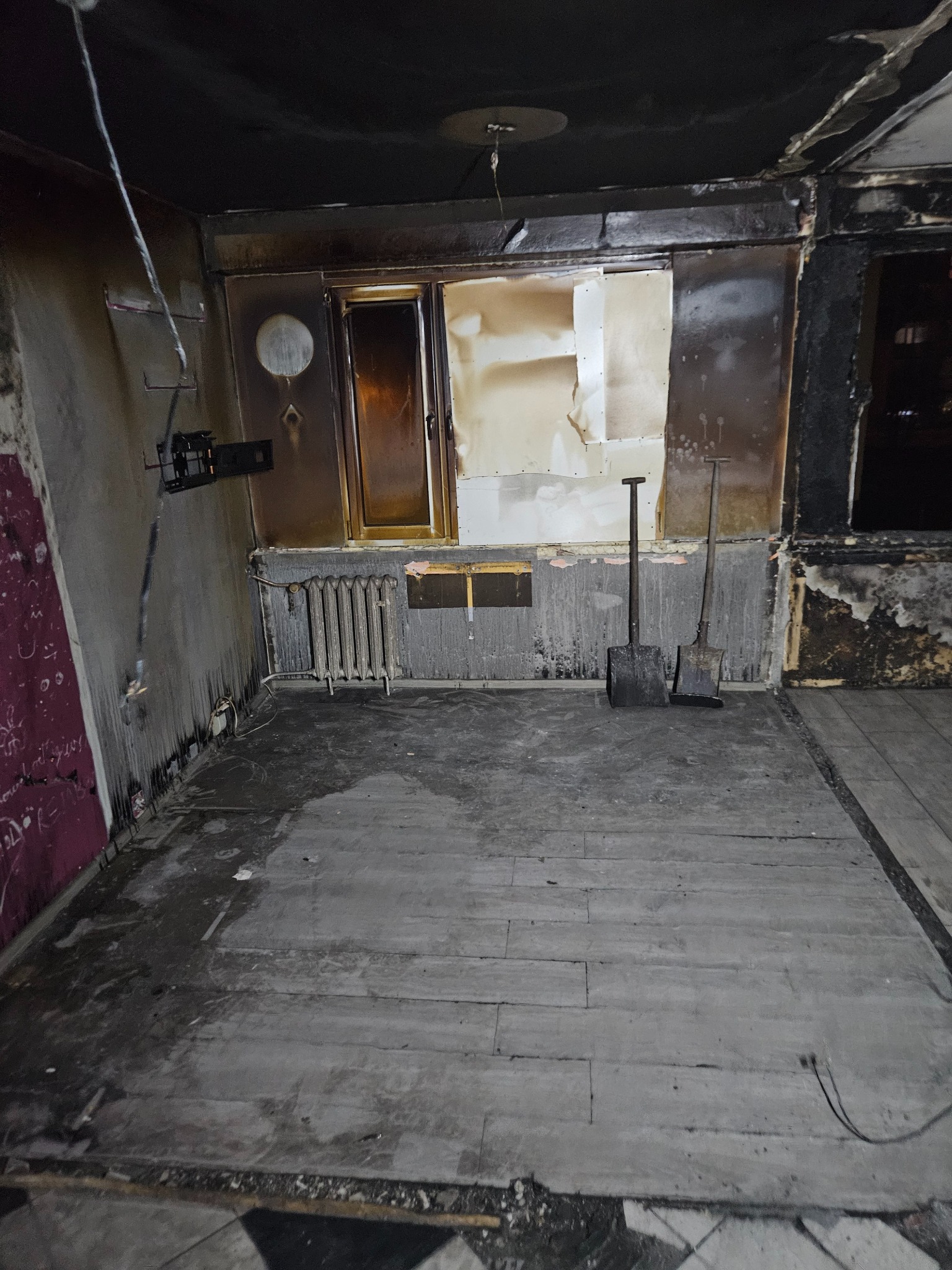 Wnętrze pokoju po pożarze z osmalonymi ścianami, oknem z częściowo usuniętą szybą, grzejnikiem, dwiema łopatami opartymi o ścianę i zniszczoną podłogą.