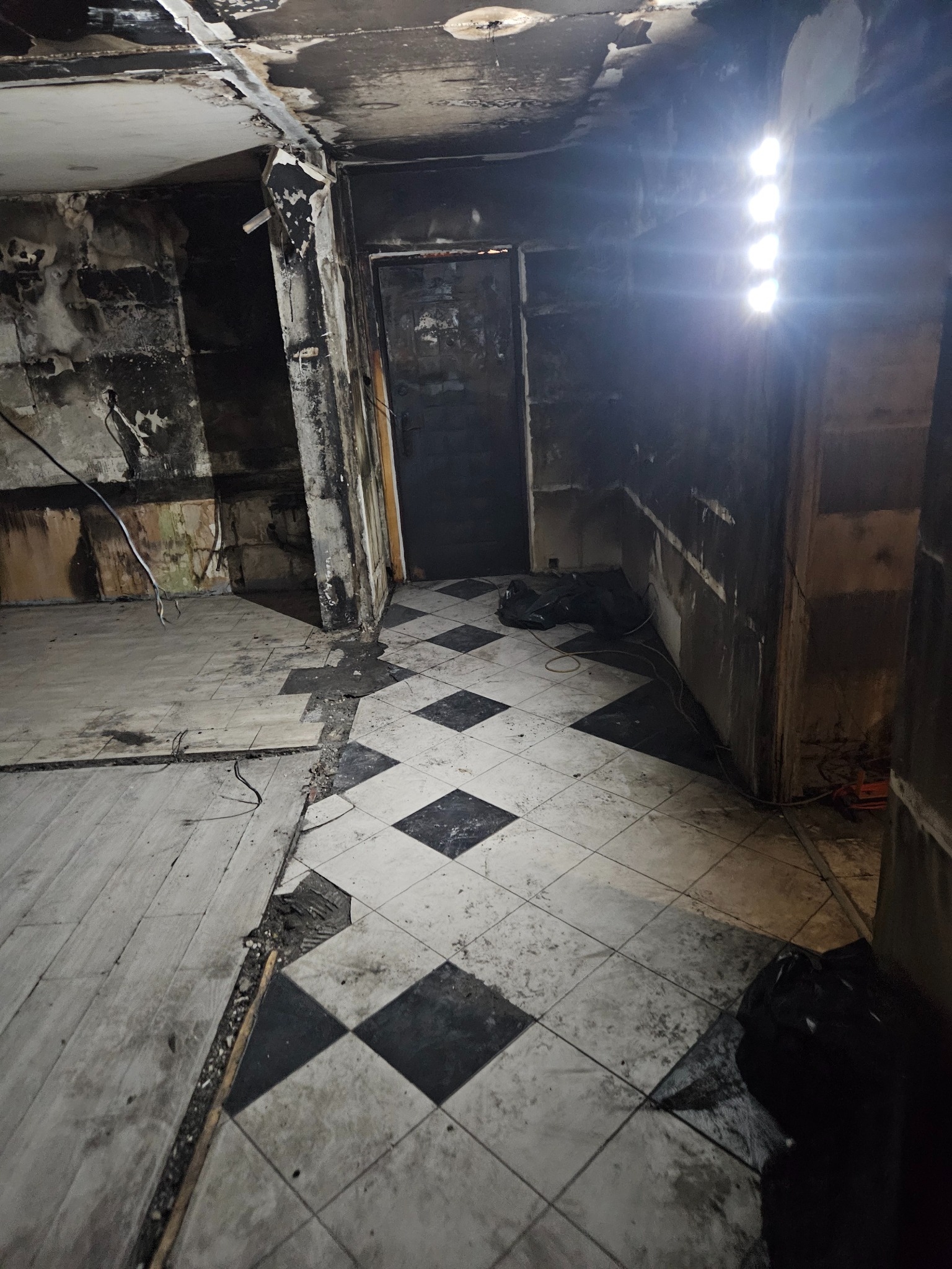 Wnętrze zniszczonego pomieszczenia z osmolonymi ścianami, uszkodzonym sufitem i podłogą w czarno-białą szachownicę; widoczne ślady po pożarze i brak wyposażenia.
