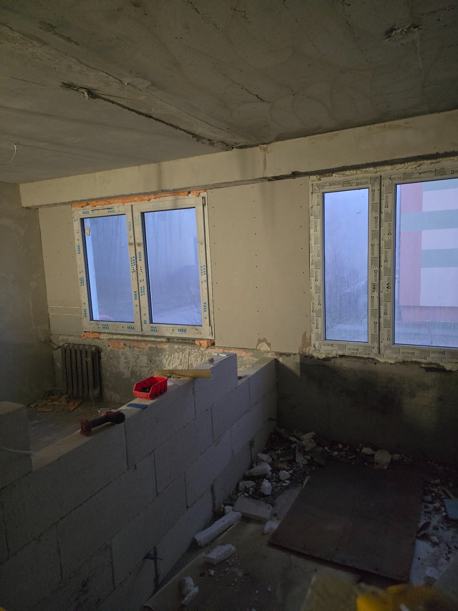 Wnętrze w trakcie prac: murowana ścianka działowa z betonu komórkowego, osadzone okna PCV z pianką montażową, surowy betonowy sufit, grzejnik żeliwny, narzędzia w czerwonym organizerze.