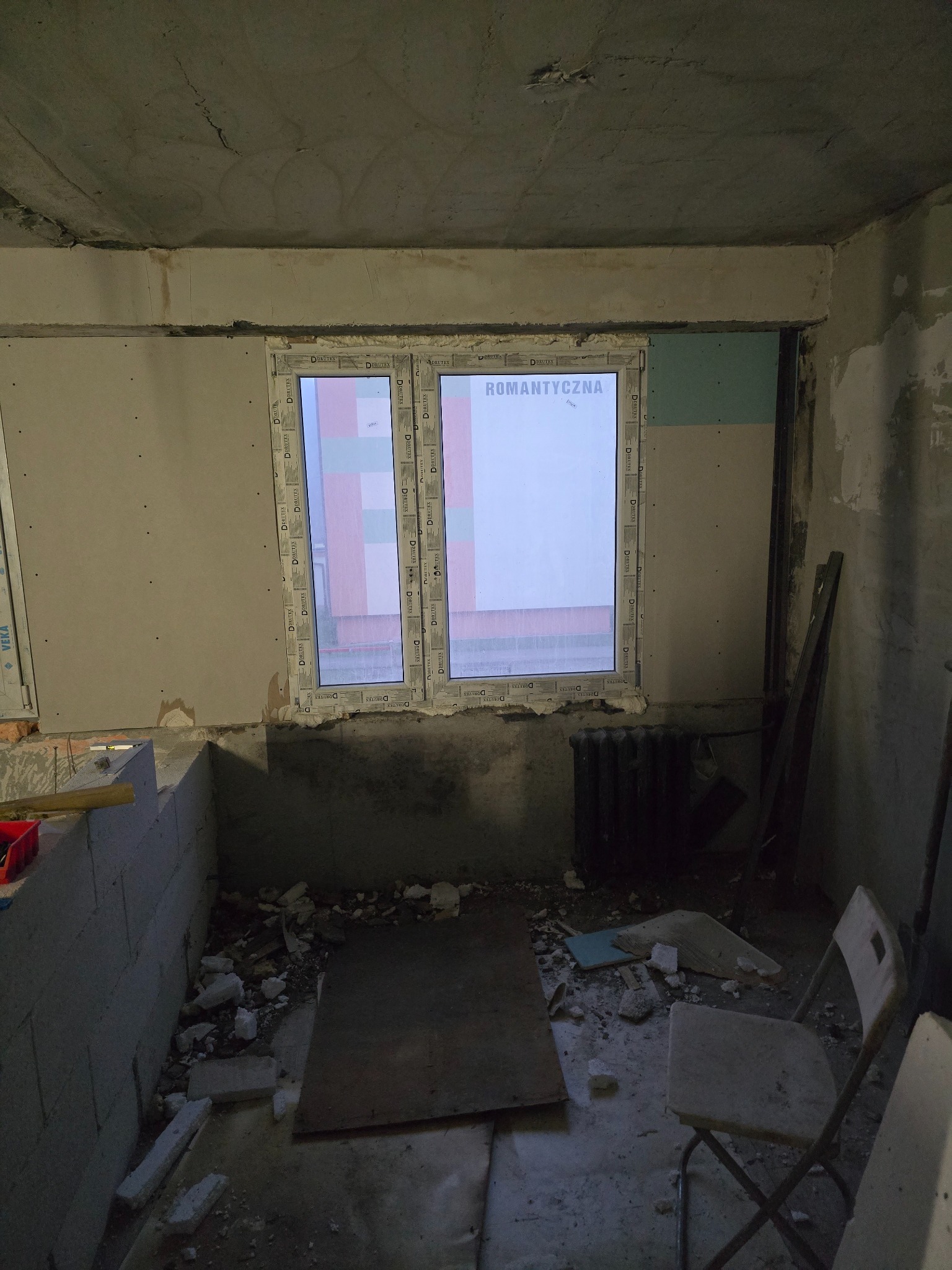 Wnętrze w trakcie prac przygotowawczych, widoczne okno z napisem 'ROMANTYCZNA', ściany z fragmentami płyt kartonowo-gipsowych, surowy strop i gruz na podłodze, krzesło z tworzywa sztucznego w rogu.