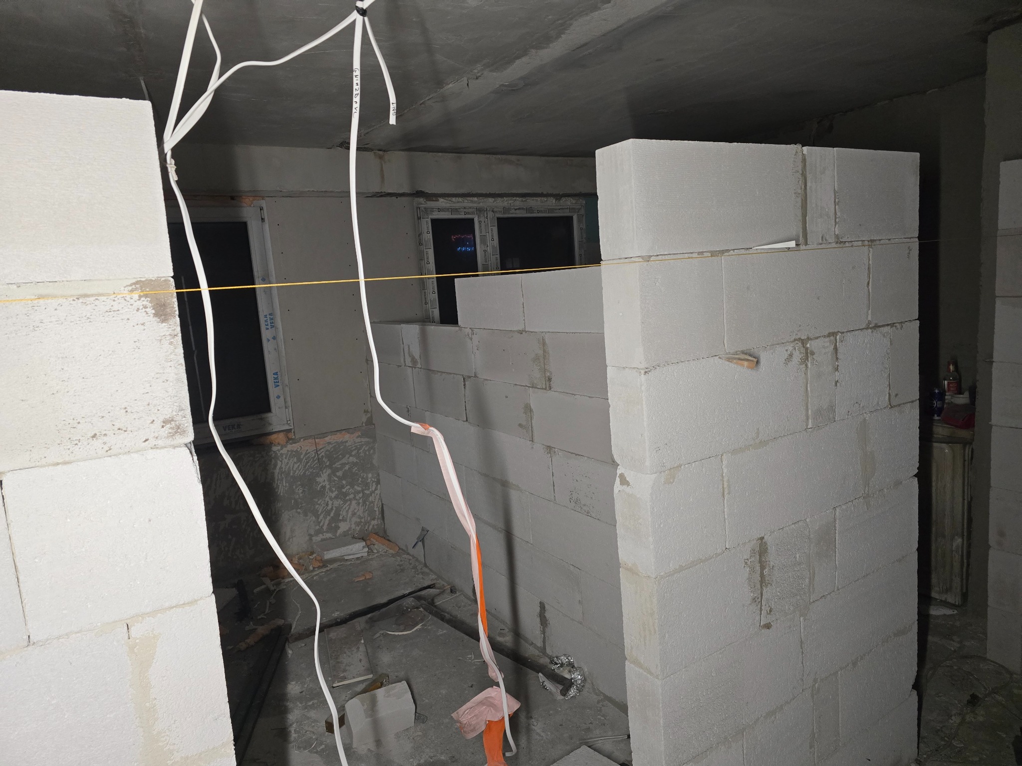 Wnętrze w trakcie budowy ścian działowych z białych bloczków, widoczne przewody elektryczne zwisające z sufitu i okno w tle.