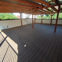 Kamil-drewniane wyroby - Zadaszony taras z desek kompozytowych w odcieniach szarości, z drewnianą konstrukcją nośną i białym murem oporowym, widok na zieleń w tle.