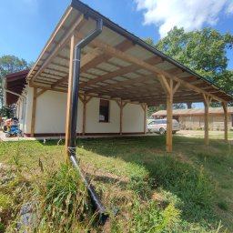 Kamil-drewniane wyroby - Drewniana konstrukcja zadaszenia, prawdopodobnie wiata garażowa, przylegająca do budynku mieszkalnego, z widocznym systemem odprowadzania wody deszczowej i samochodem w tle.