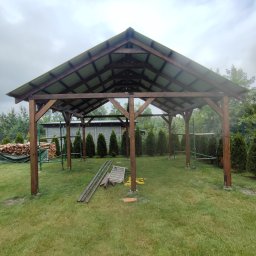 Kamil-drewniane wyroby - Drewniana konstrukcja z zadaszeniem w trakcie budowy, widoczne rusztowania i materiały budowlane na trawniku, pochmurne niebo w tle.
