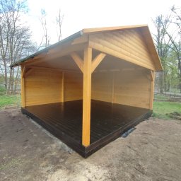 Kamil-drewniane wyroby - Nowo wybudowana, drewniana konstrukcja z dachem, prawdopodobnie altana lub wiata, z pomalowaną na ciemno podłogą, stojąca na ziemi.