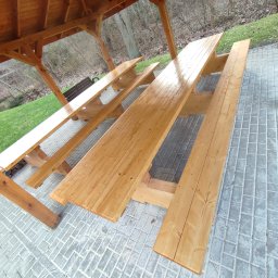 Kamil-drewniane wyroby - Nowa, drewniana konstrukcja z ławami i stołami piknikowymi pod zadaszeniem, widok z góry na szarej kostce brukowej, trawa i drzewa w tle.