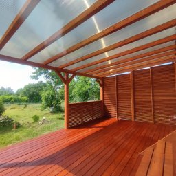 Kamil-drewniane wyroby - Zadaszony taras z drewnianą podłogą w kolorze terakoty, częściowo osłonięty drewnianymi panelami, z widokiem na zielony ogród.