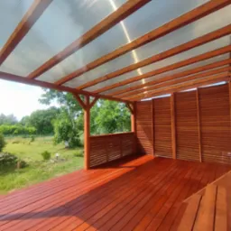 Zadaszony taras z drewnianą podłogą w kolorze terakoty, częściowo osłonięty drewnianymi panelami, z widokiem na zielony ogród.