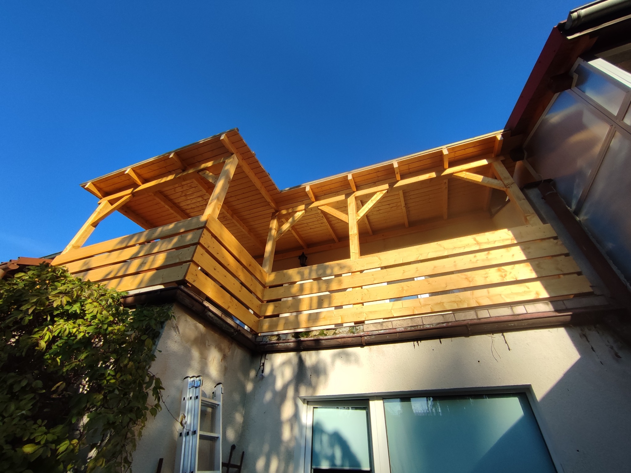 Drewniana konstrukcja z belek i desek, prawdopodobnie zadaszenie lub balkon, w trakcie budowy, widok z dołu na tle błękitnego nieba, drabina oparta o ścianę budynku w pobliżu.