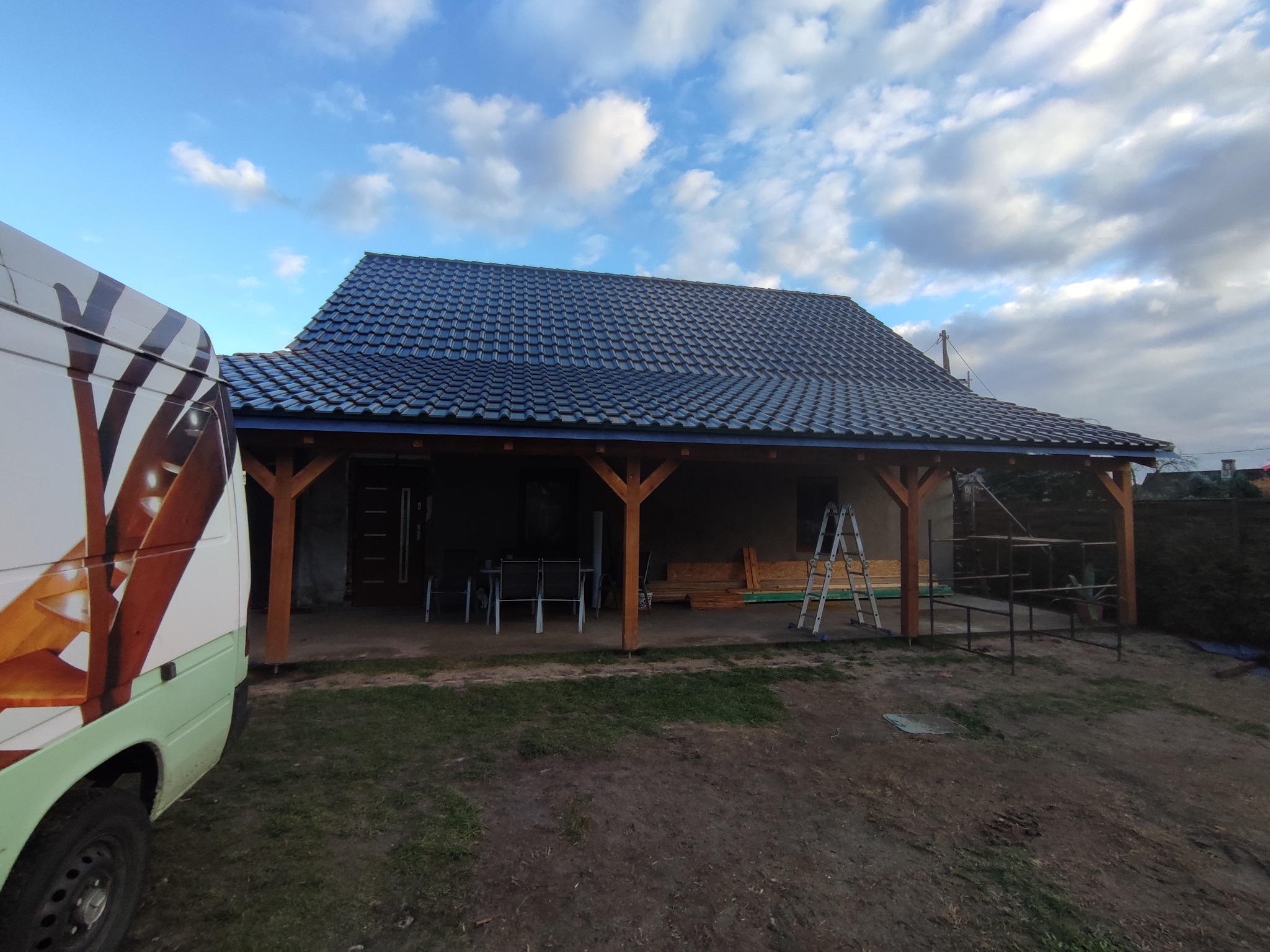 Zadaszony taras z ciemną dachówką, wsparty na drewnianych słupach, widok z zewnątrz, z boku zaparkowany samochód z grafiką schodów.