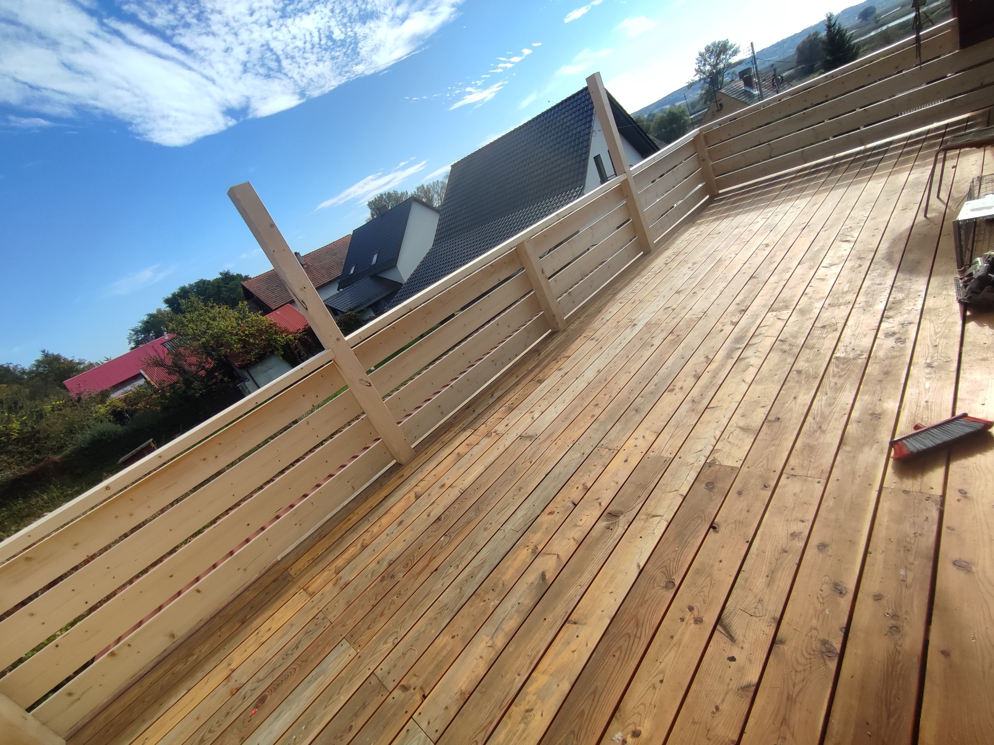 Nowo wybudowany taras z jasnego drewna z drewnianą balustradą, widok na domy i błękitne niebo z chmurami, na podłodze leży szczotka.