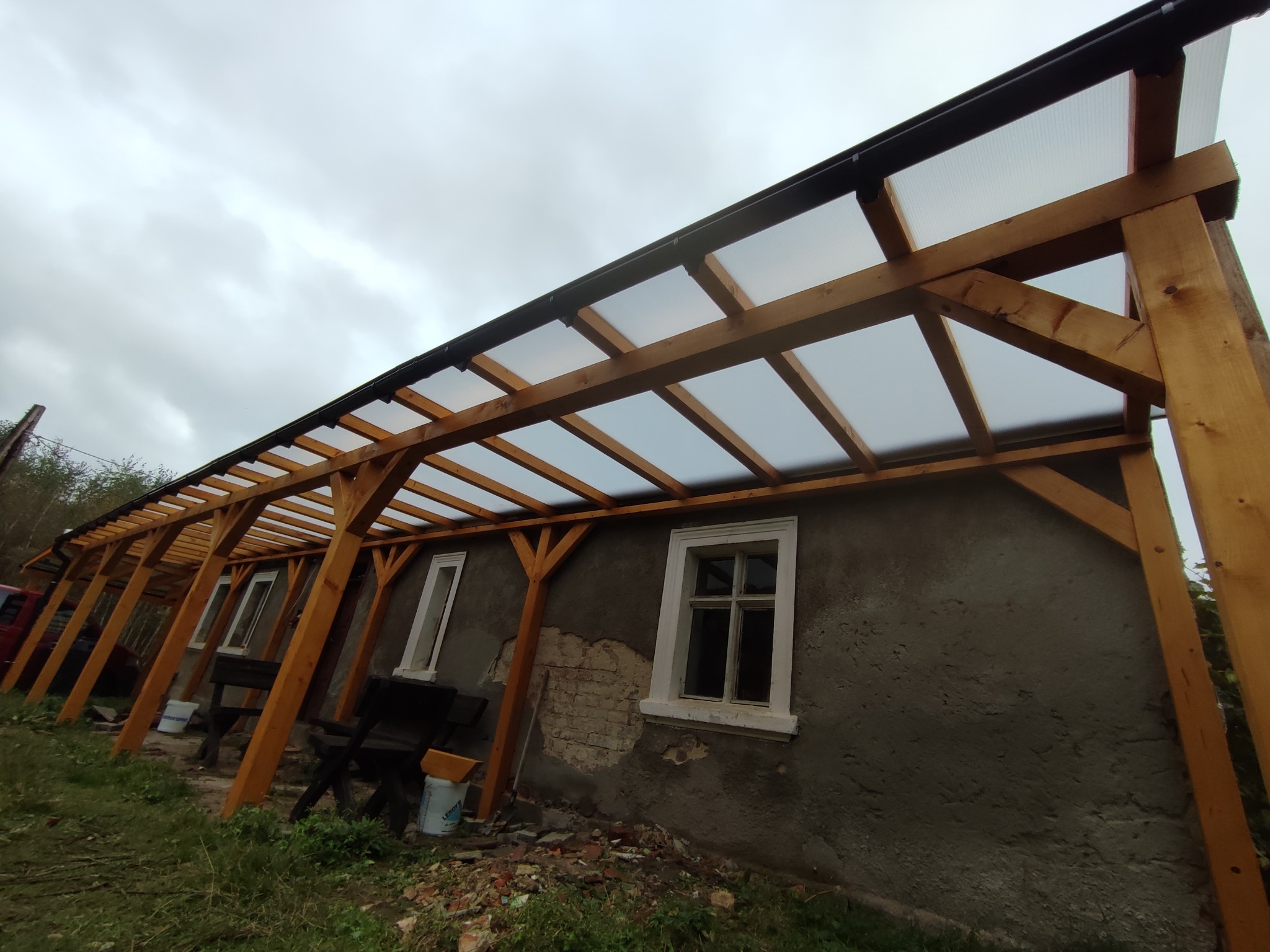 Zadaszenie z drewna i przezroczystych paneli przy szarym budynku z białymi oknami, widok z dołu w pochmurny dzień.