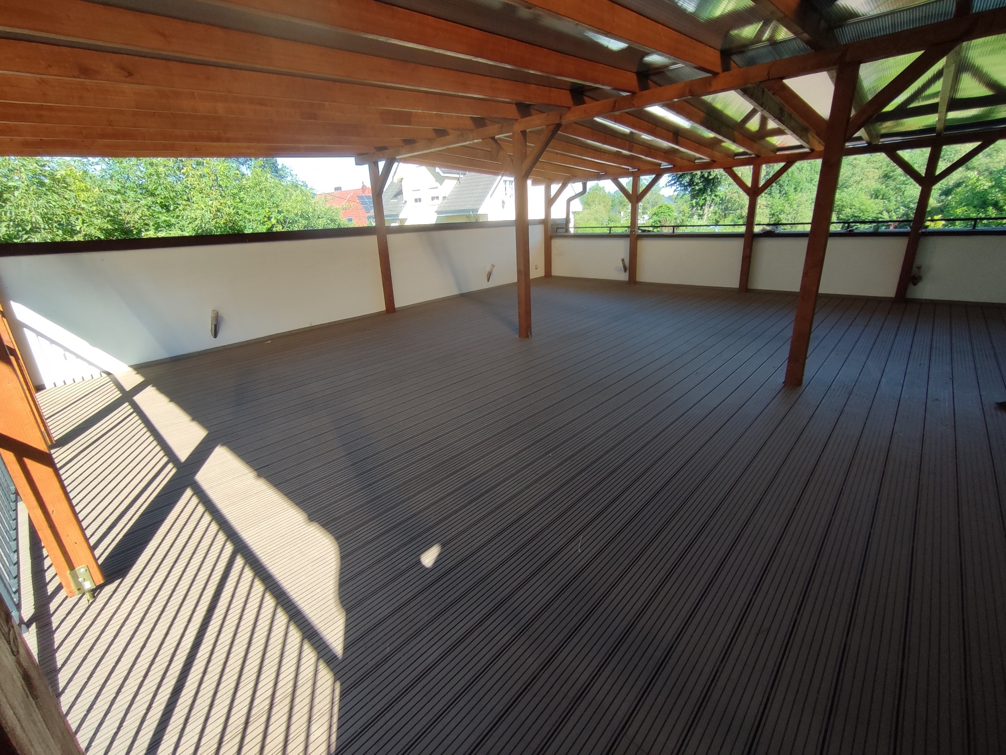 Zadaszony taras z desek kompozytowych w odcieniach szarości, z drewnianą konstrukcją nośną i białym murem oporowym, widok na zieleń w tle.