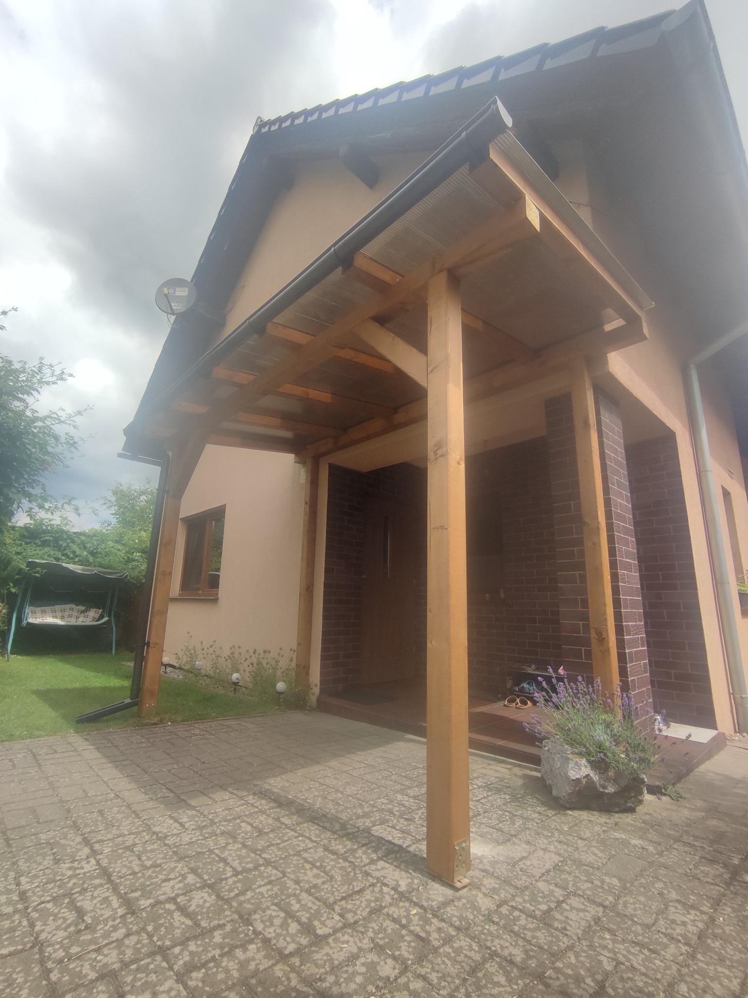 Drewniana wiata przy wejściu do domu, z przeszklonym dachem, obok donica z lawendą i widoczny fragment brukowanej ścieżki.