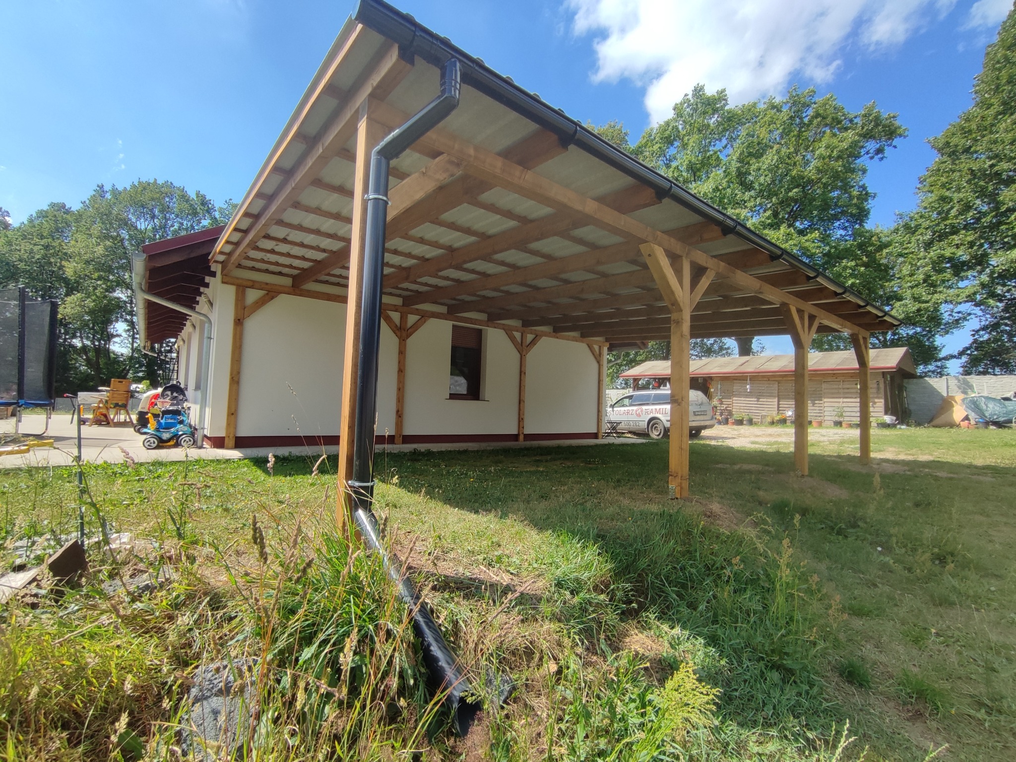 Drewniana konstrukcja zadaszenia, prawdopodobnie wiata garażowa, przylegająca do budynku mieszkalnego, z widocznym systemem odprowadzania wody deszczowej i samochodem w tle.