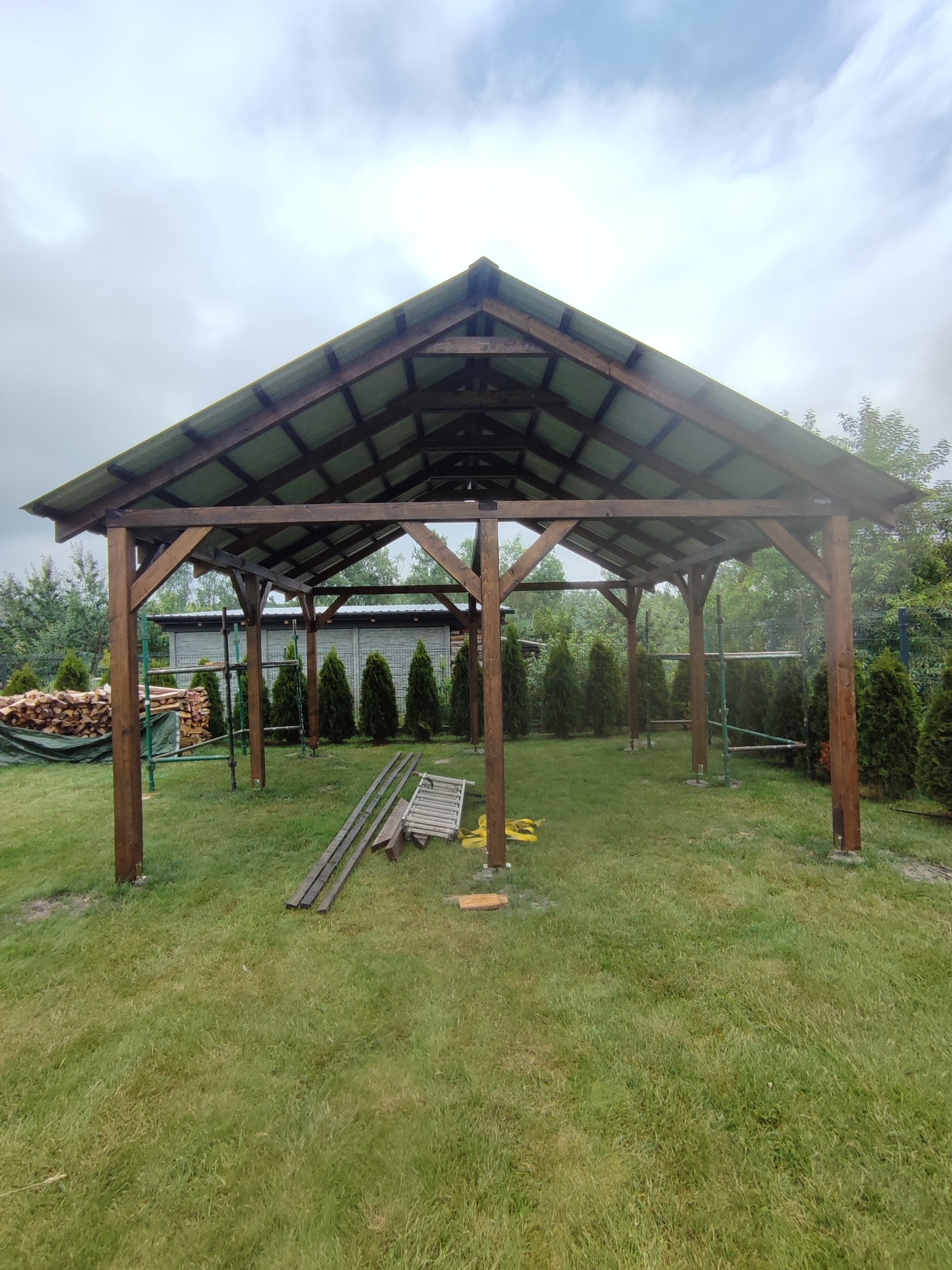 Drewniana konstrukcja z zadaszeniem w trakcie budowy, widoczne rusztowania i materiały budowlane na trawniku, pochmurne niebo w tle.