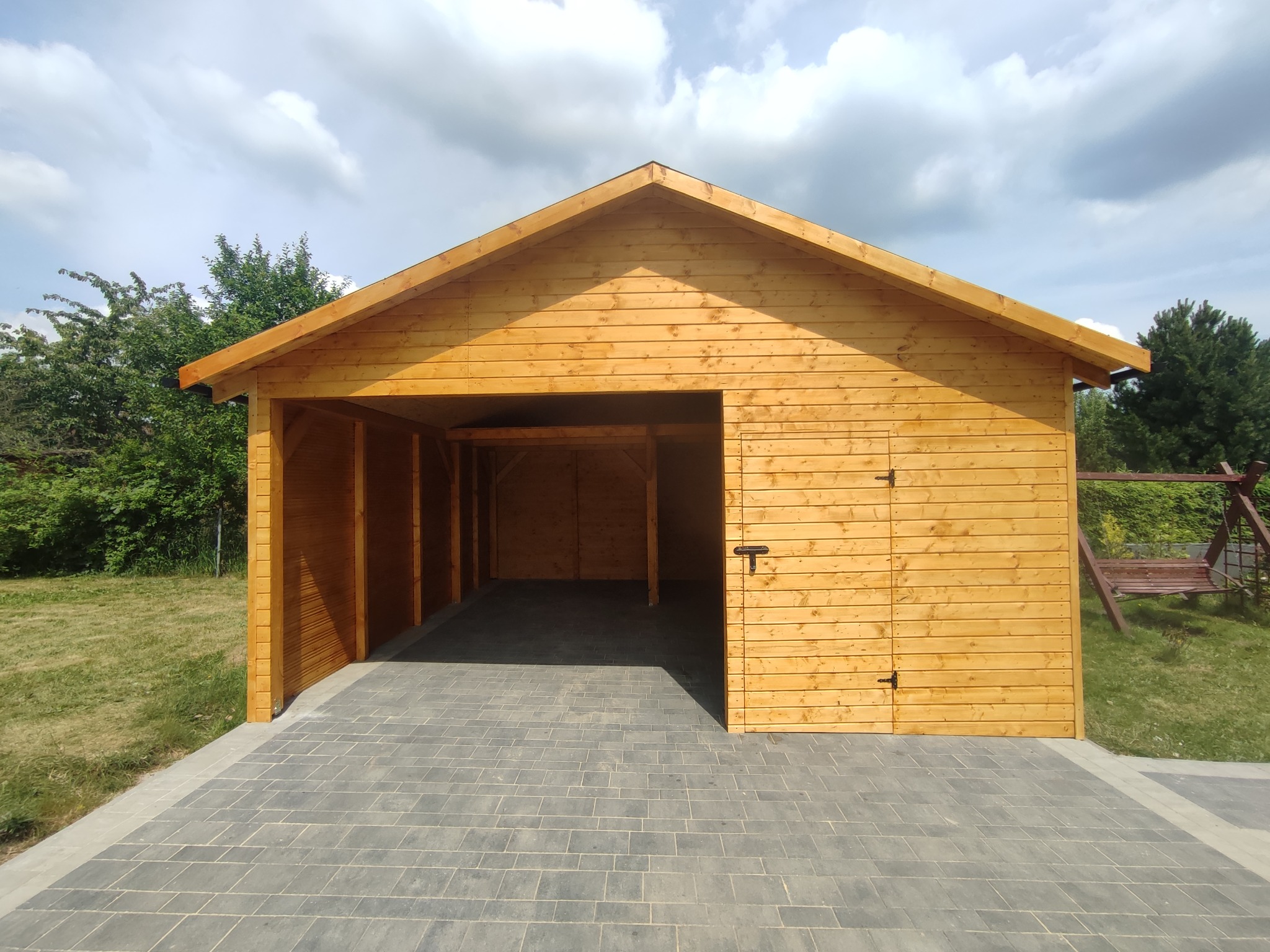 Drewniana altana z zamykaną częścią, świeżo po impregnacji, na wyłożonym kostką podłożu, w tle zieleń.
