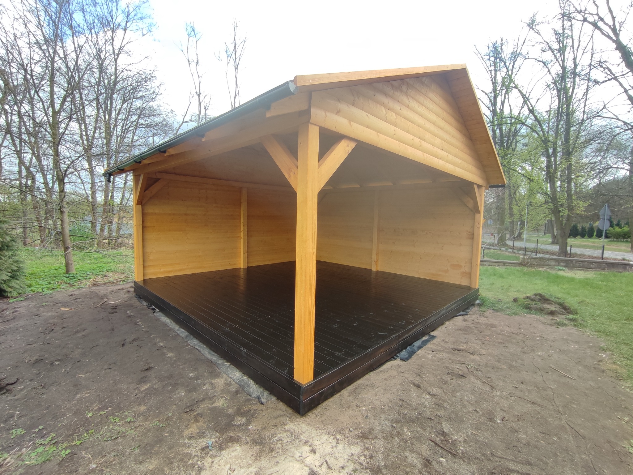 Nowo wybudowana, drewniana konstrukcja z dachem, prawdopodobnie altana lub wiata, z pomalowaną na ciemno podłogą, stojąca na ziemi.