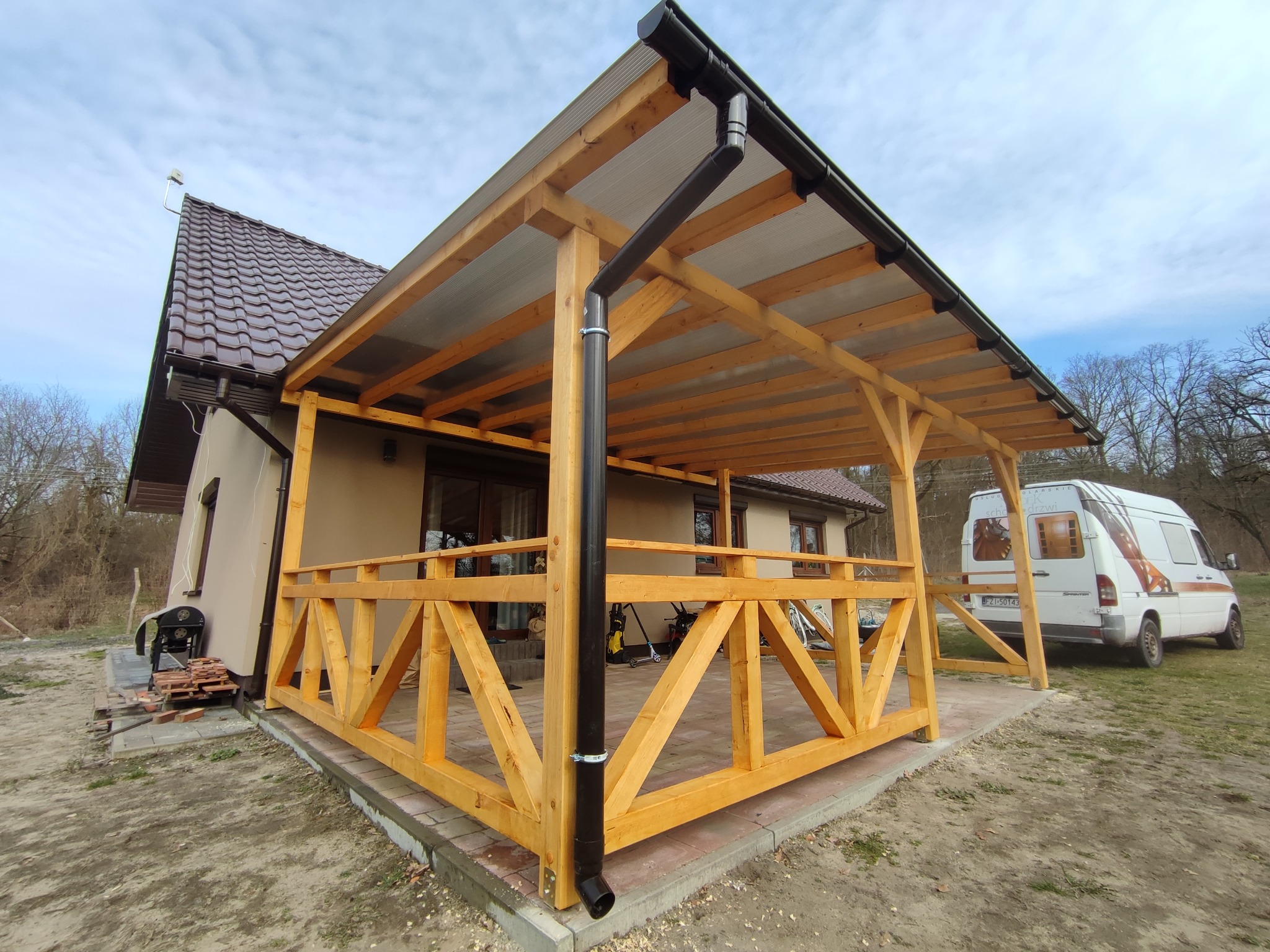 Surowa drewniana konstrukcja zadaszonej przestrzeni, prawdopodobnie w trakcie budowy, przylegająca do domu w Bobrowicach, z widocznym systemem rynnowym i stertą płytek na ziemi.