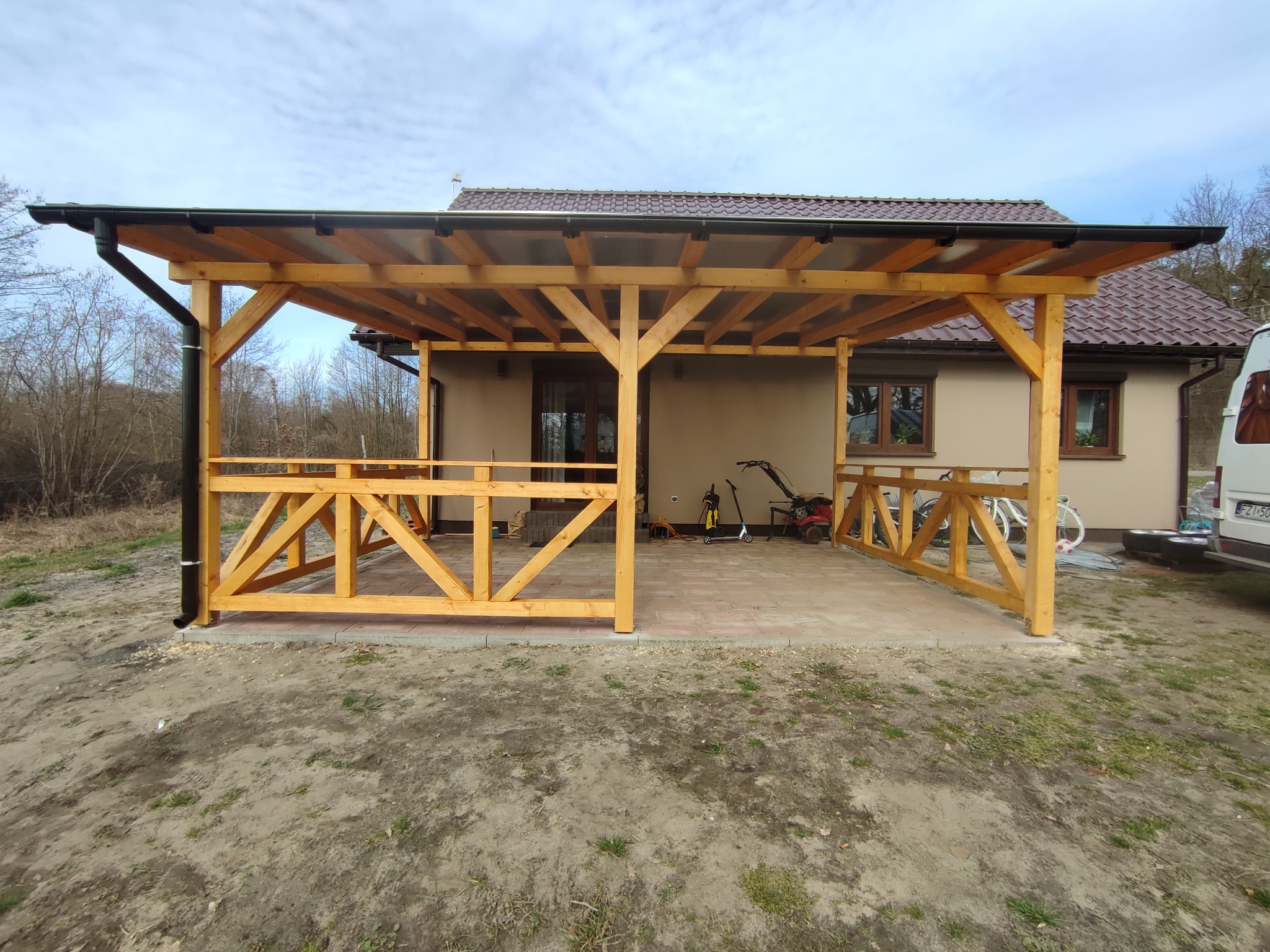 Surowa drewniana konstrukcja z przezroczystym zadaszeniem, prawdopodobnie w trakcie budowy, z widocznymi narzędziami i sprzętem ogrodniczym w tle, na częściowo wyłożonym kostką brukową terenie.