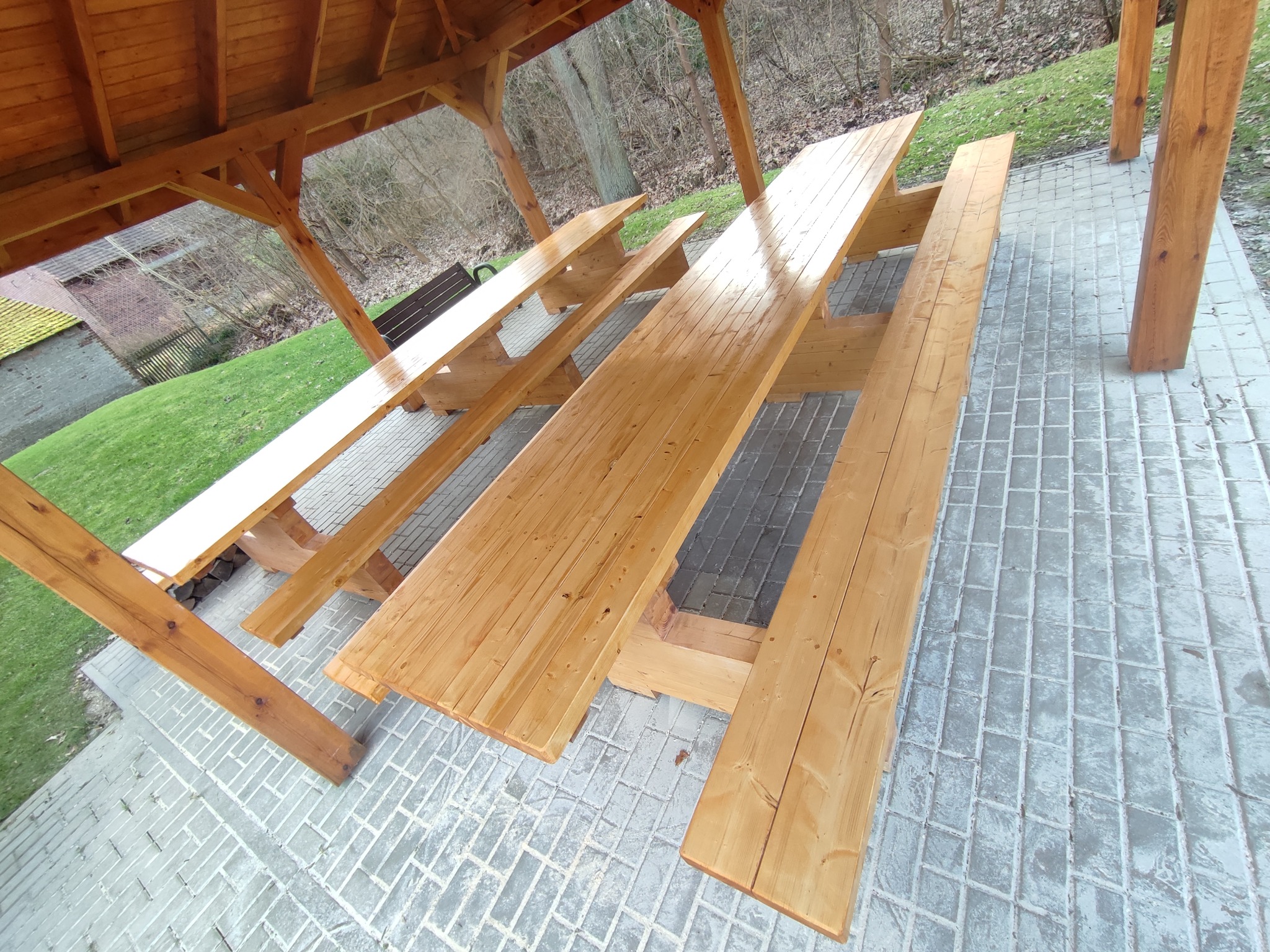 Nowa, drewniana konstrukcja z ławami i stołami piknikowymi pod zadaszeniem, widok z góry na szarej kostce brukowej, trawa i drzewa w tle.