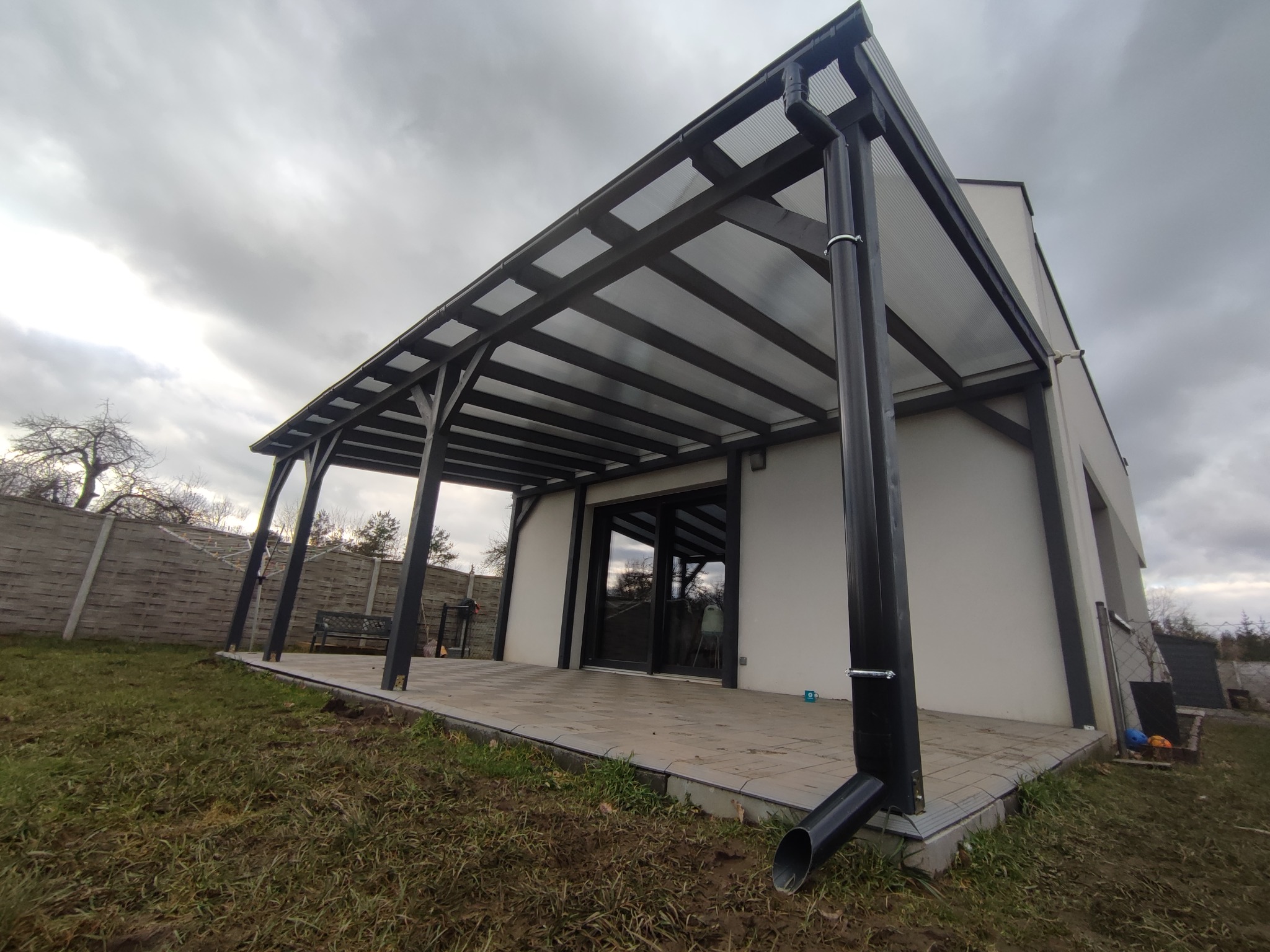 Nowoczesna pergola z ciemną konstrukcją i przezroczystym zadaszeniem, przylegająca do budynku z tarasem wyłożonym kostką, widok na pochmurne niebo.