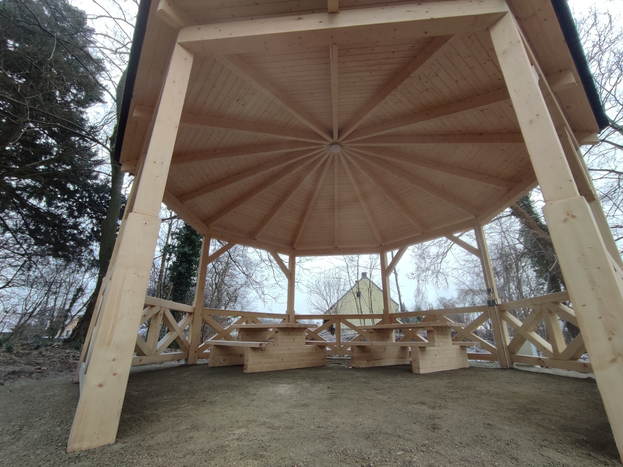 Ośmiokątna altana z surowego drewna z wbudowanymi ławkami i stołami, widok od dołu ukazujący konstrukcję dachu i oświetlenie, w tle drzewa i dom.