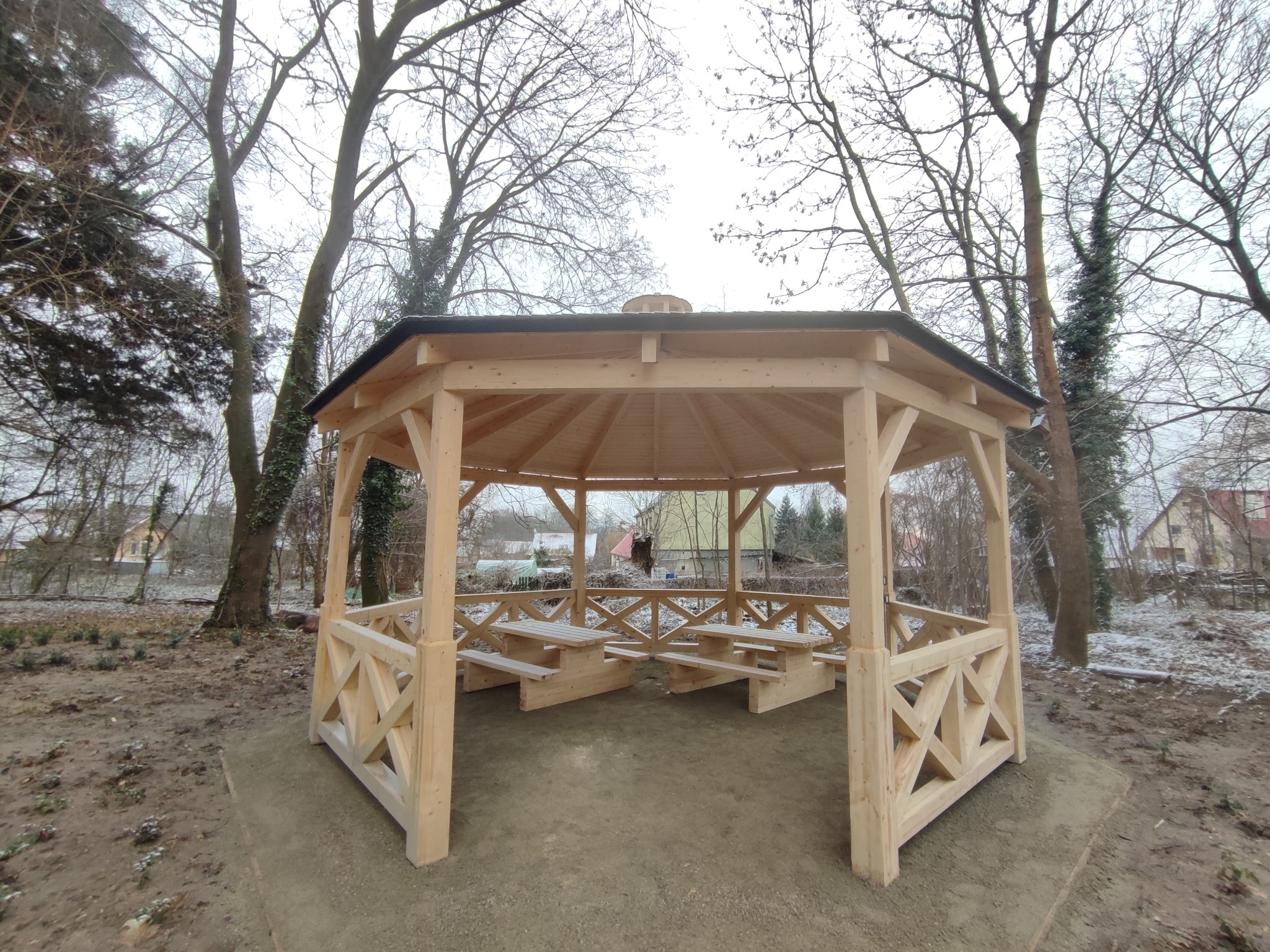Ośmioboczna, drewniana konstrukcja z zadaszeniem i wbudowanymi ławkami, otoczona drzewami w zimowej scenerii.