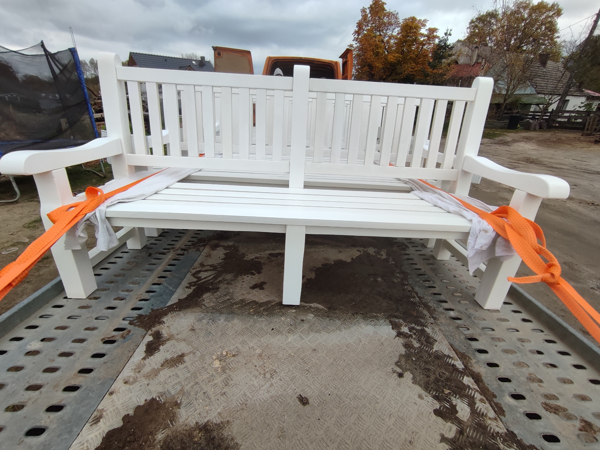 Nowa, biała, drewniana ławka ogrodowa zabezpieczona pomarańczowymi pasami transportowymi na przyczepie samochodowej.