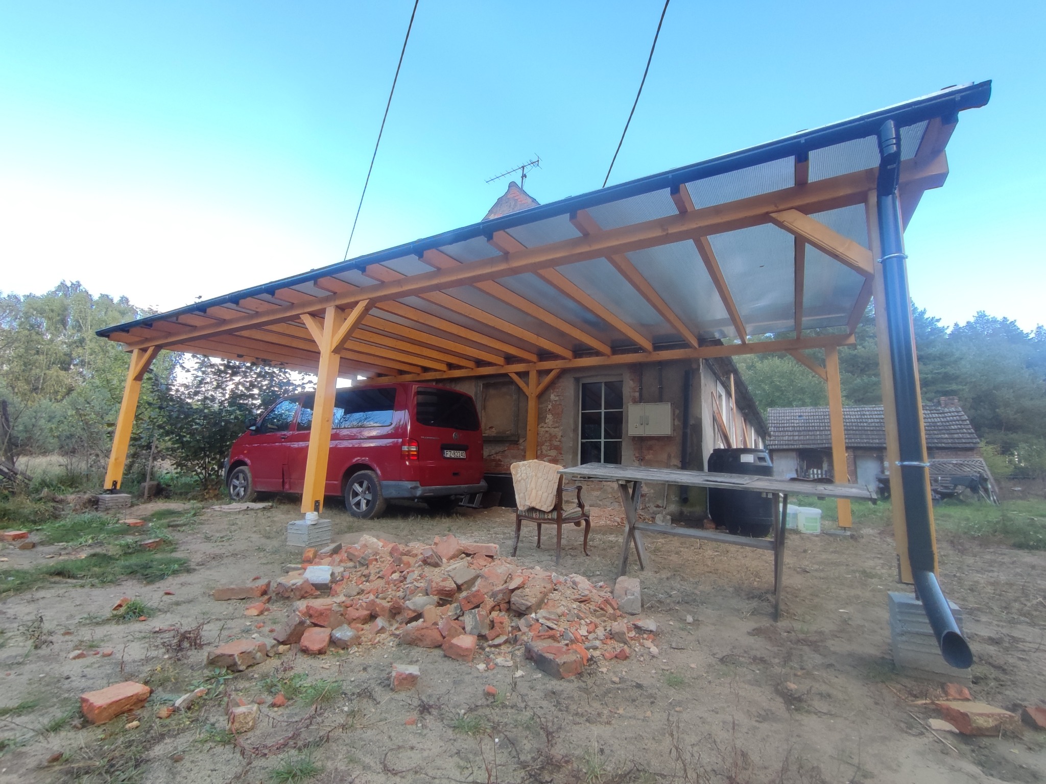 Nowa drewniana konstrukcja z przezroczystym zadaszeniem, prawdopodobnie wiata garażowa, z widocznym samochodem osobowym w tle, starym budynkiem i stertą gruzu z cegieł na pierwszym planie.