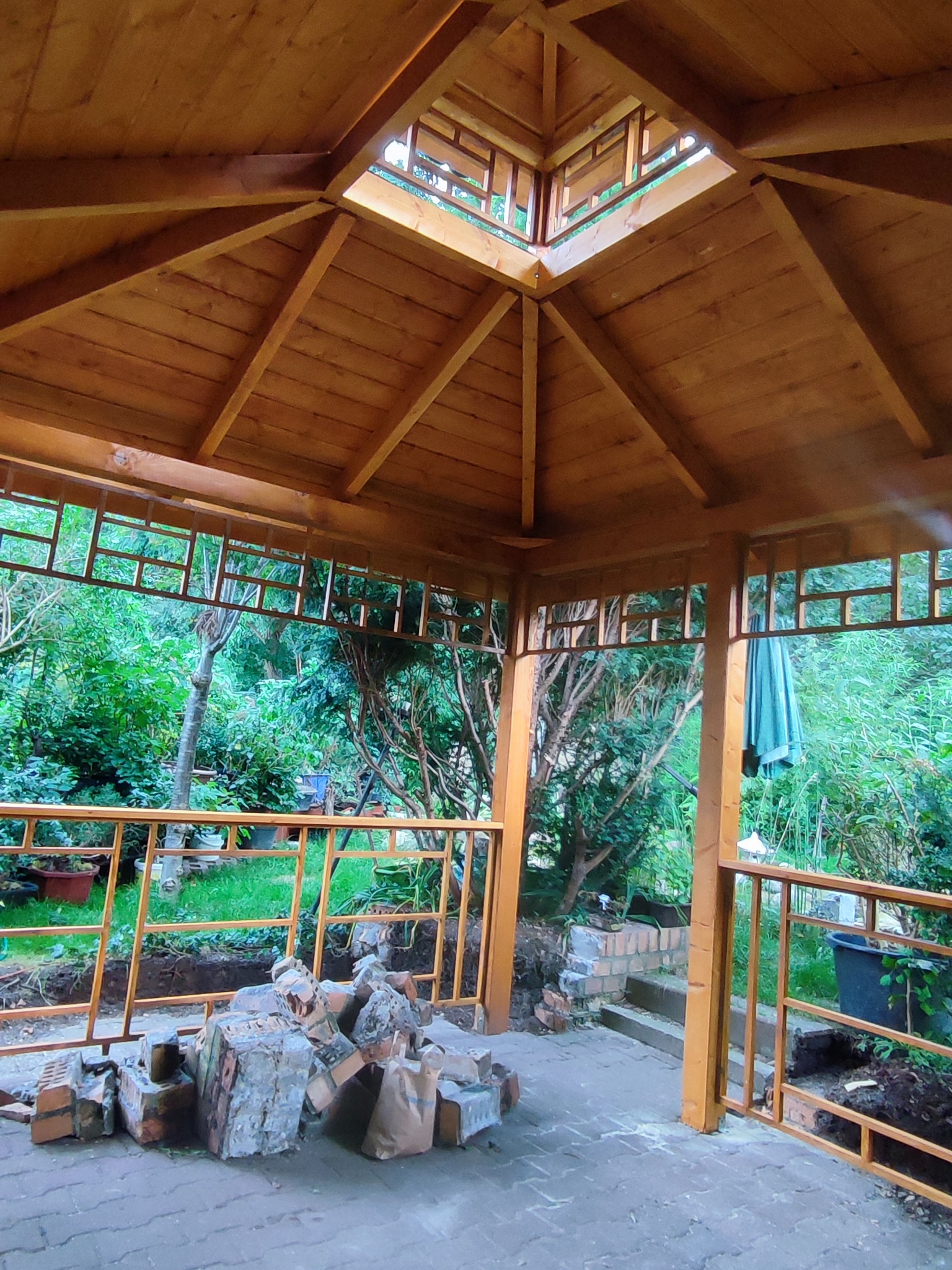 Wnętrze drewnianej konstrukcji z widocznym dachem i ażurowymi ścianami, w tle bujna zieleń ogrodu, a na pierwszym planie cegły i materiały budowlane sugerujące trwające prace wykończeniowe.