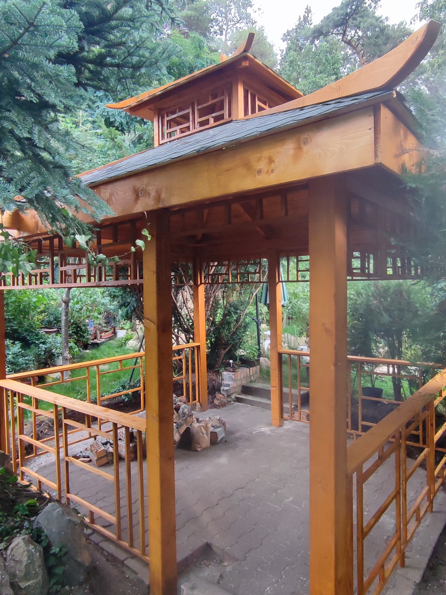 Drewniana konstrukcja w stylu pagody z charakterystycznym zakrzywionym dachem, otoczona zielenią ogrodu, z widocznymi elementami niedokończonej budowy wewnątrz.