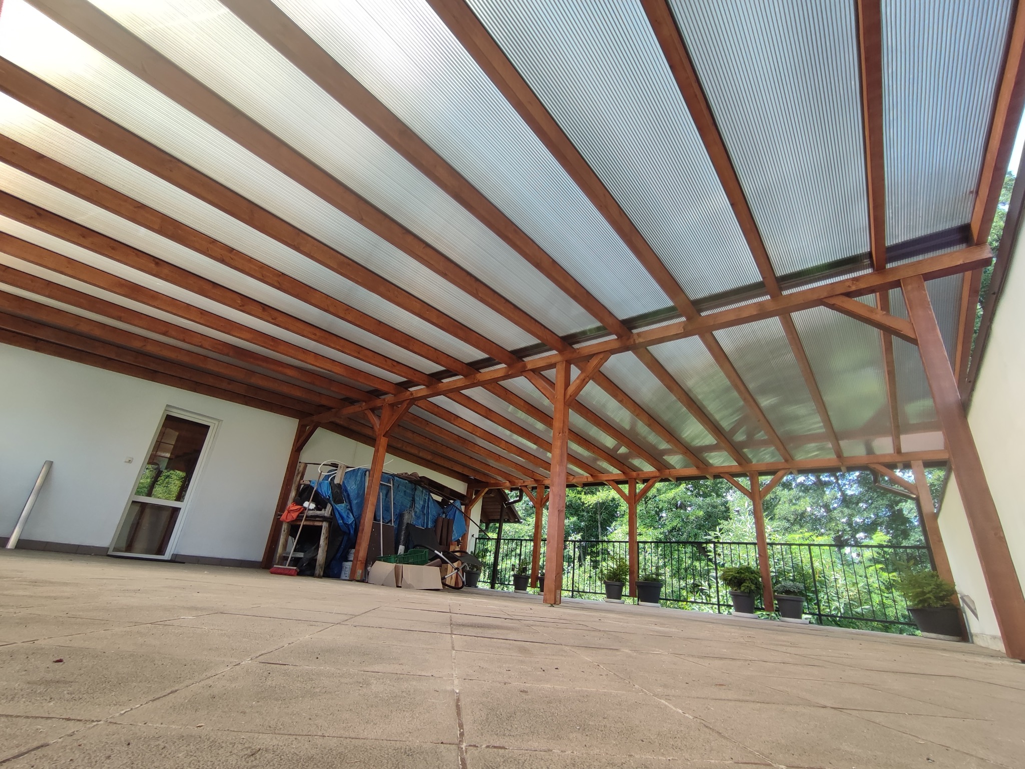 Zadaszenie tarasu z drewnianą konstrukcją i przezroczystym poliwęglanowym dachem, widok z dołu na zewnątrz, z widocznymi roślinami w donicach na tarasie.