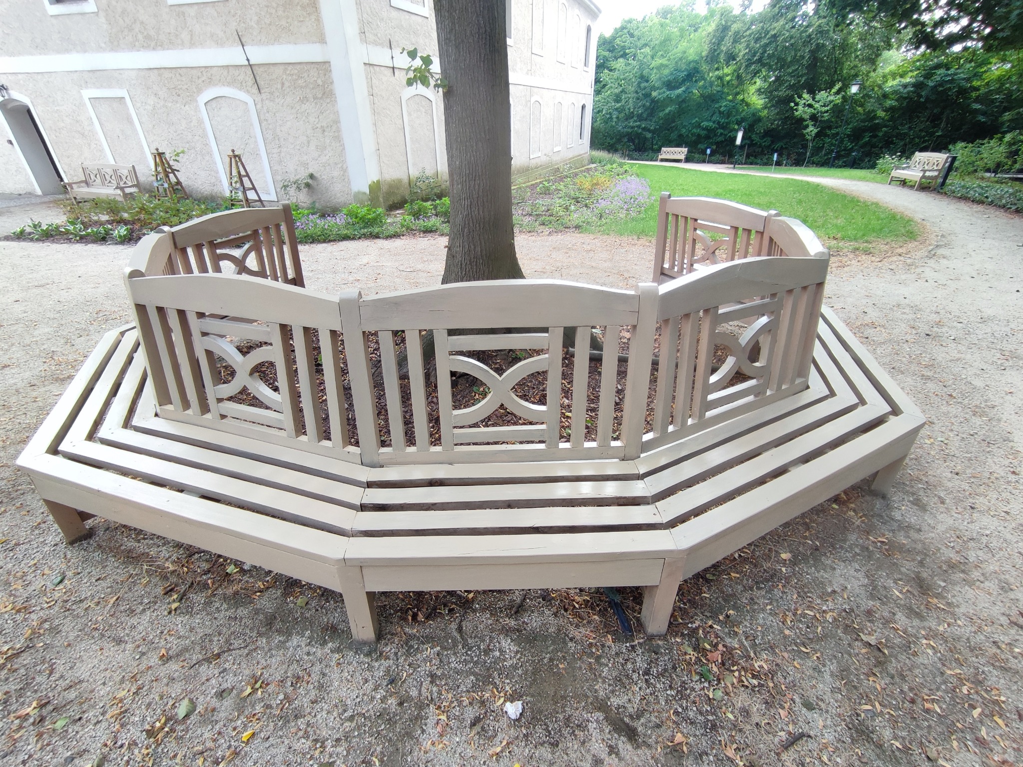 Półokrągła ławka z drewna otaczająca pień drzewa w parku, z widocznymi elementami dekoracyjnymi i budynkiem w tle.