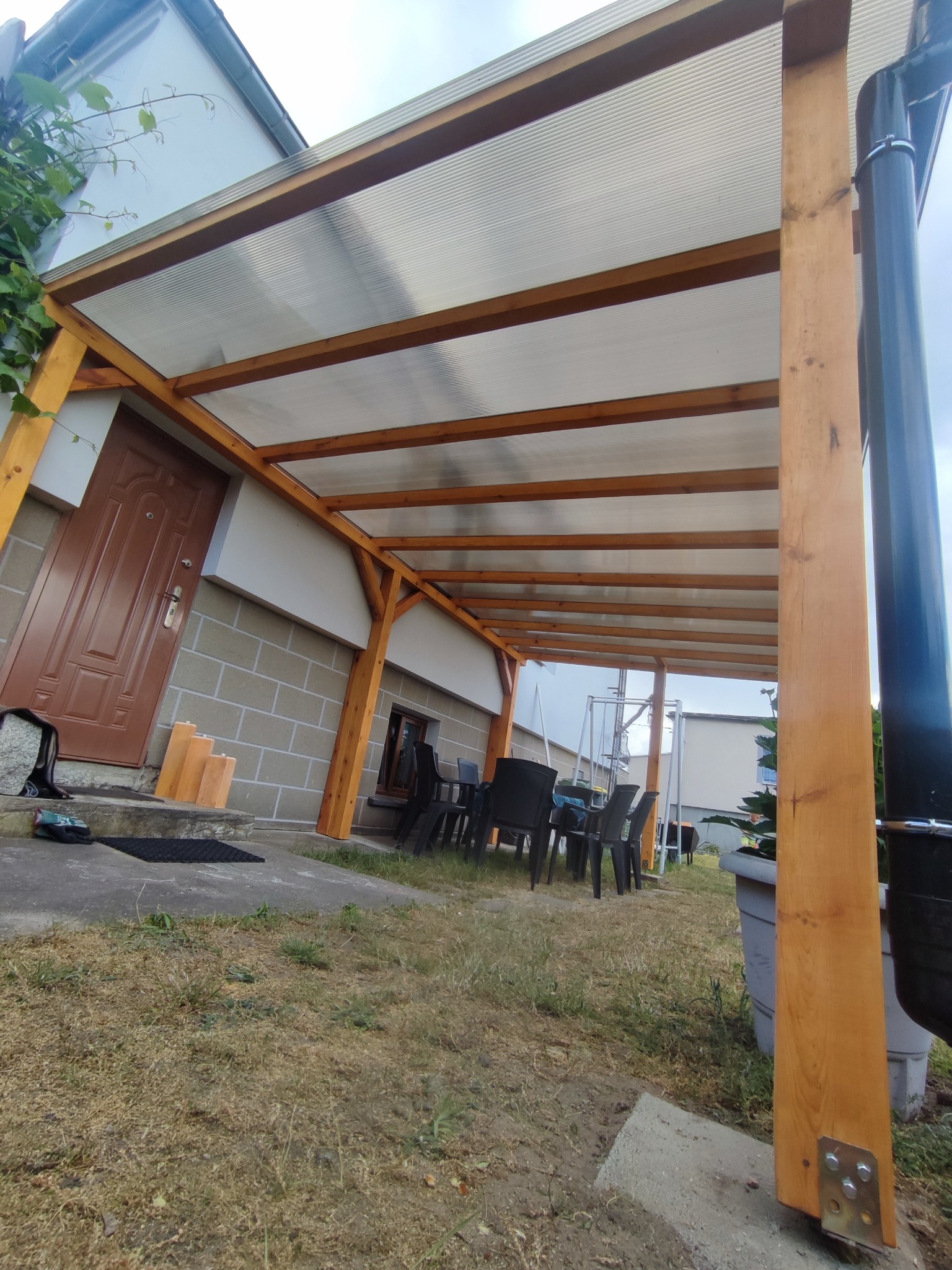 Drewniana pergola z przezroczystym zadaszeniem z poliwęglanu, przylegająca do ściany domu, z widocznymi elementami mocującymi i zestawem mebli ogrodowych w tle.