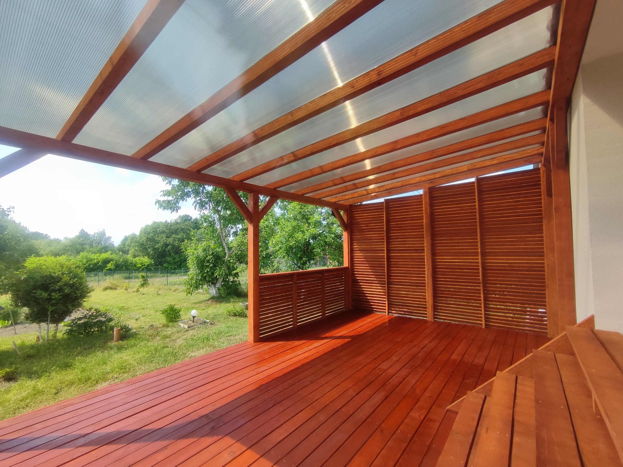 Zadaszony taras z drewnianą podłogą w kolorze terakoty, częściowo osłonięty drewnianymi panelami, z widokiem na zielony ogród.