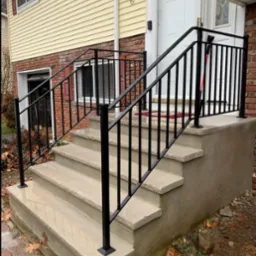 Nowoczesna, czarna balustrada ze stali na betonowych schodach wejściowych do domu z żółtą elewacją i ceglanym cokołem, widok z dołu.