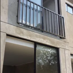 Nowoczesna, minimalistyczna balustrada balkonowa z pionowych, metalowych prętów, zamontowana na surowej, betonowej elewacji budynku z dużymi przesuwnymi oknami.