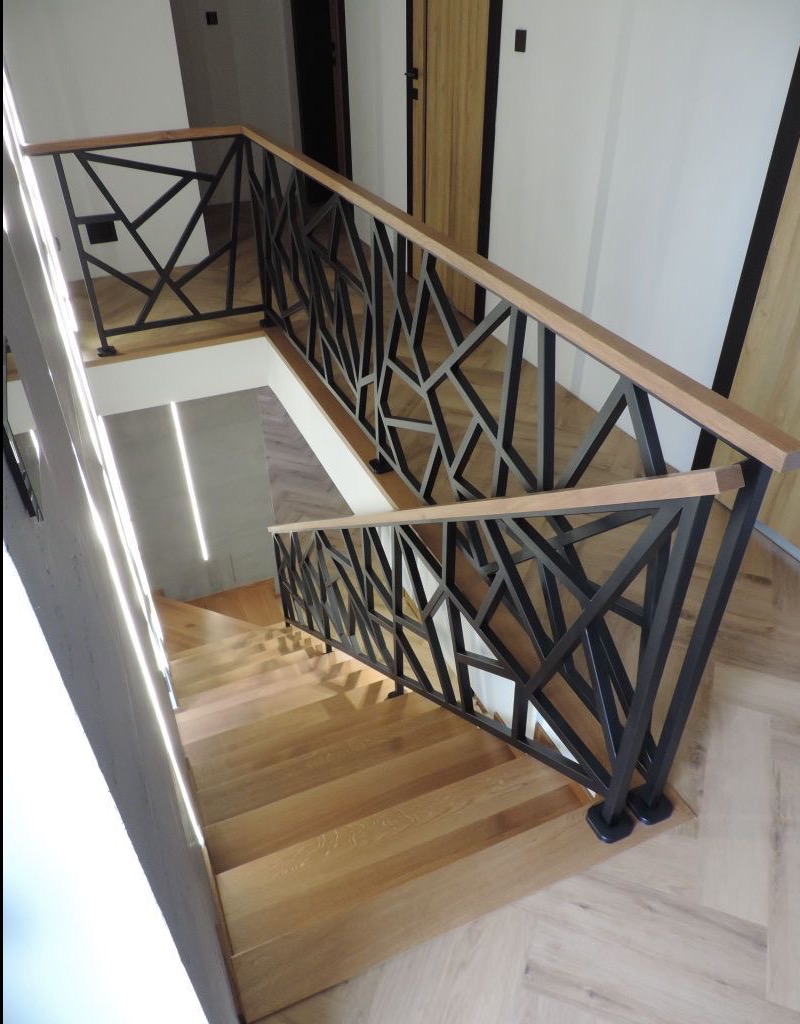 Nowoczesna balustrada schodowa z czarnych, geometrycznych elementów i drewnianym poręczą, widok z góry na jasne drewniane schody i parkiet ułożony w jodełkę.