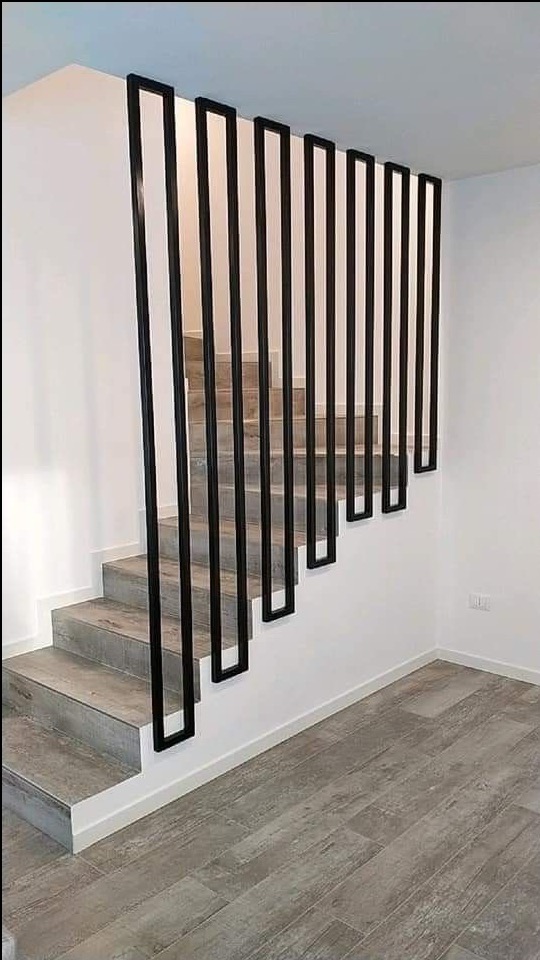 Nowoczesna balustrada schodowa z czarnych, pionowych profili o kwadratowym przekroju, mocowanych do ściany i stopni, w minimalistycznym wnętrzu z szarymi płytkami podłogowymi i schodami.