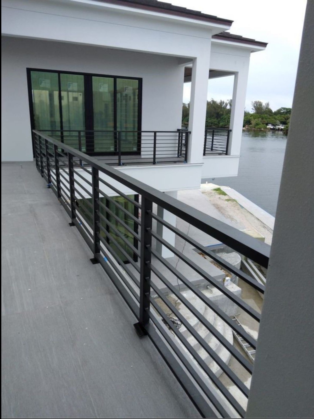 Nowoczesna, czarna balustrada metalowa z poziomymi prętami na balkonie w Łomiankach, z widokiem na wodę i budynek w tle.
