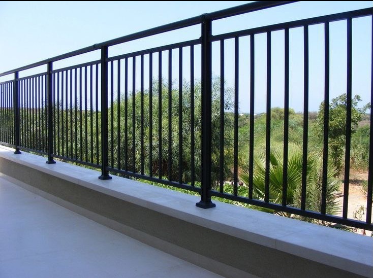 Czarna, metalowa balustrada balkonowa z widokiem na bujną roślinność i błękitne niebo w tle.