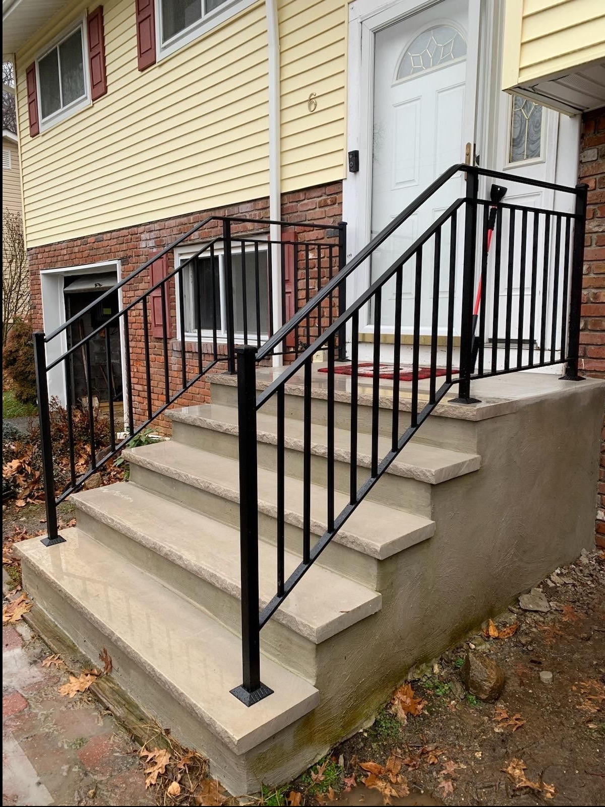 Nowoczesna, czarna balustrada ze stali na betonowych schodach wejściowych do domu z żółtą elewacją i ceglanym cokołem, widok z dołu.
