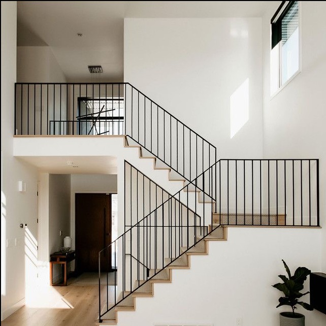 Nowoczesne wnętrze z białymi ścianami i schodami z czarną, minimalistyczną balustradą, widok na antresolę z podobną balustradą i drewnianą podłogą, roślina doniczkowa w rogu.